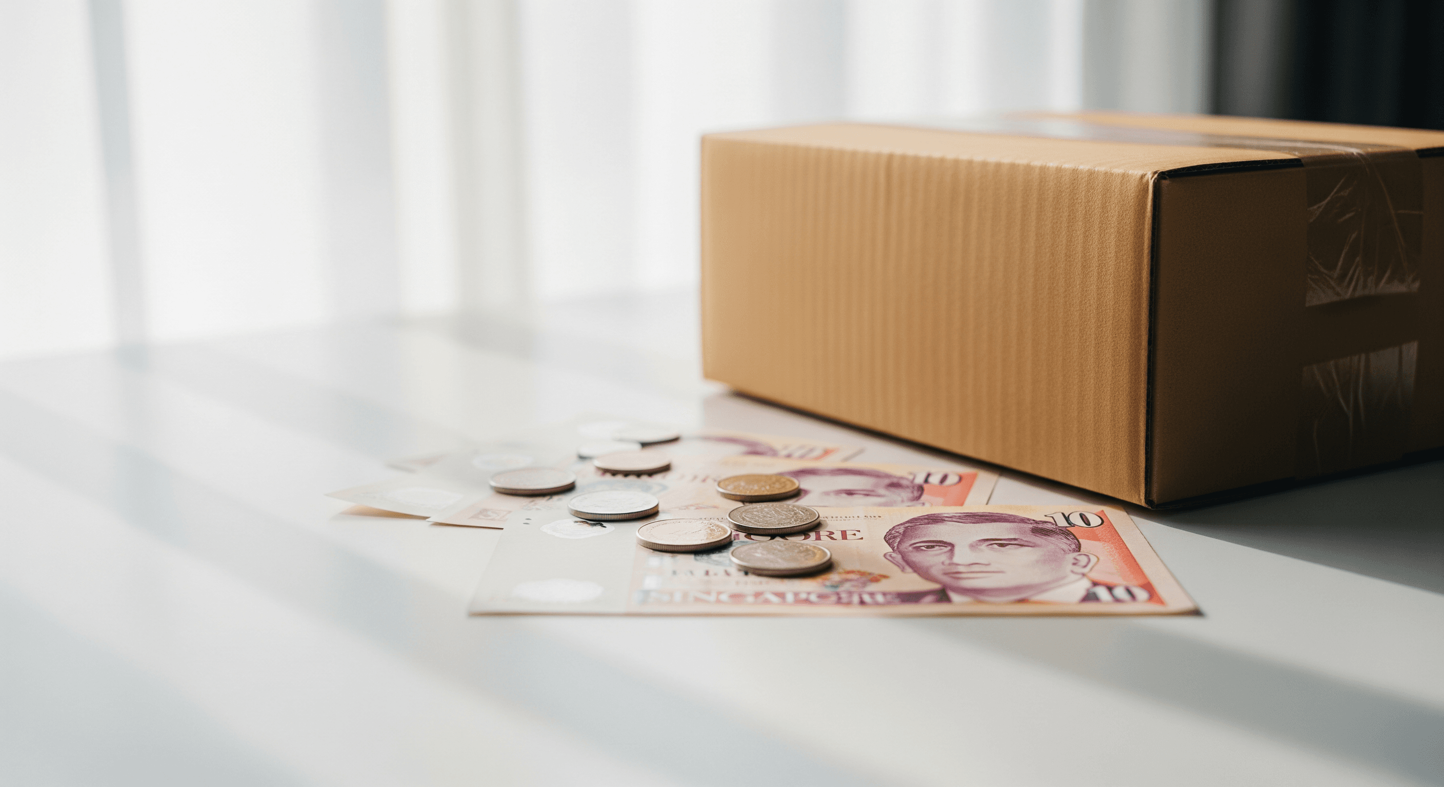 Singapore dollar notes and coins next to a cardboard delivery parcel on a white desk